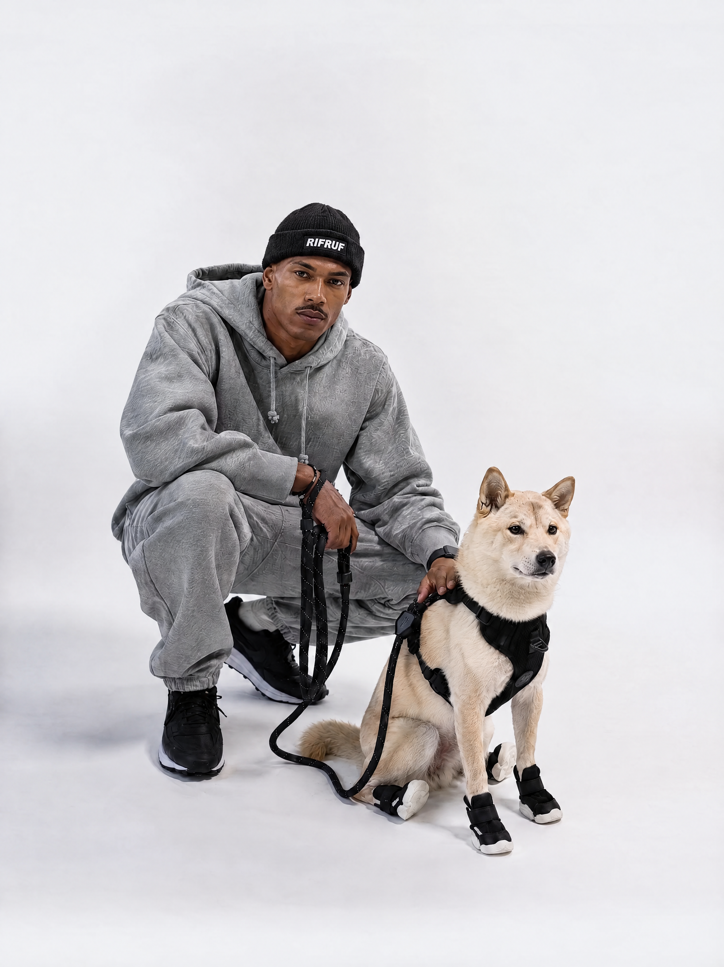Model wearing the black RIFRUF Beanie, crouching beside a Shiba Inu in a black RIFRUF harness, leash and dog shoes